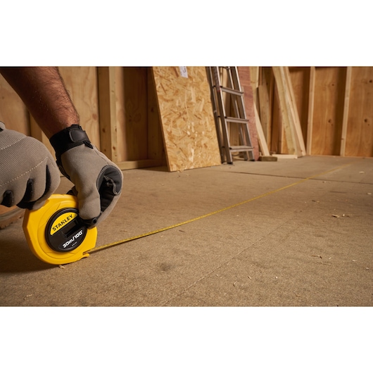 STANLEY 0-34-107 measuring tape in use on construction floor, gloves holding yellow case.