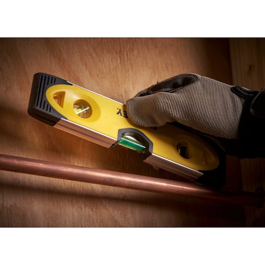 STANLEY 0-43-511 spirit level used by a gloved hand to check copper pipe alignment against a wooden surface.