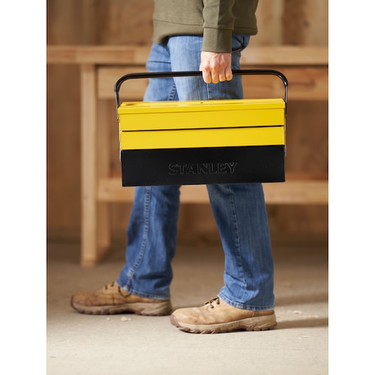 Stanley 1-94-738 yellow and black metal cantilever toolbox being carried by a person, indoor setting.