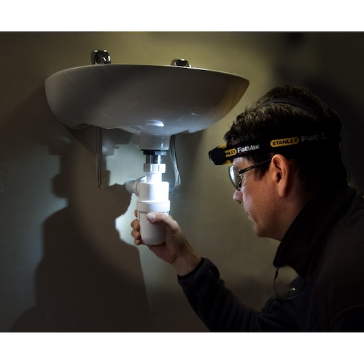 STANLEY FMHT0-70767 headlamp used by a person working under a sink, illuminating plumbing in a dark setting.