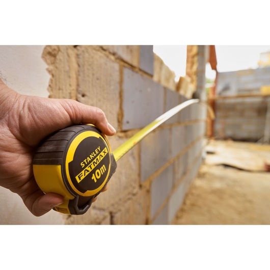 Photo of STANLEY FMHT33010-3 FATMAX 10m tape measure held against a brick wall at a construction site.
