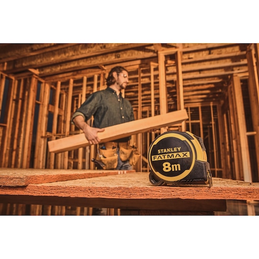 STANLEY FMHT33103-3 FATMAX 8m tape measure on wood, construction site background with worker holding timber.
