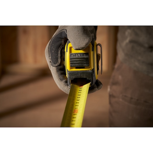 Photo of STANLEY FMHT33103 tape measure held by gloved hand, yellow and black body with extended tape.