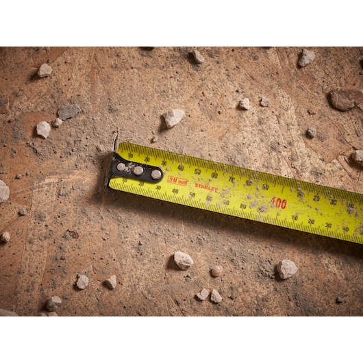 Photo of STANLEY FMHT33103 tape measure extended on rough, dusty ground with small rocks.