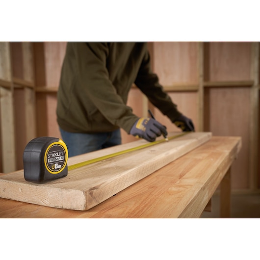 STANLEY FMHT33869 tape measure on wood plank with person marking measurements in workshop.