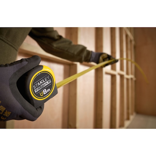 Stanley FMHT33869 tape measure with yellow blade extended, held by gloved hand at construction framing.