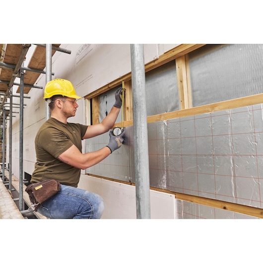 Photo of STANLEY FMHT38218-5 tape measure in use by a construction worker measuring insulation on a building wall.