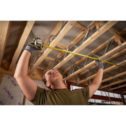 STANLEY FMHT38218-5 tape measure used by person wearing gloves to measure wooden ceiling beams in construction site.