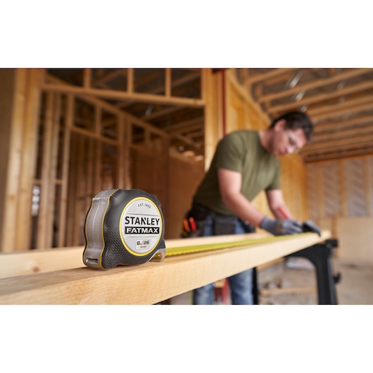 Photo of a STANLEY FATMAX tape measure, SKU FMHT38218-5, on a wooden beam at a construction site, with a worker in the background.