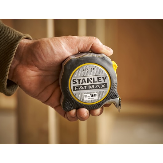 Stanley FMHT38218-5 FatMax tape measure held in hand, black and yellow casing, indoor setting.