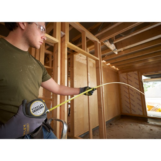 STANLEY FMHT38233-5 tape measure extended by worker in gloves at a wooden construction site.