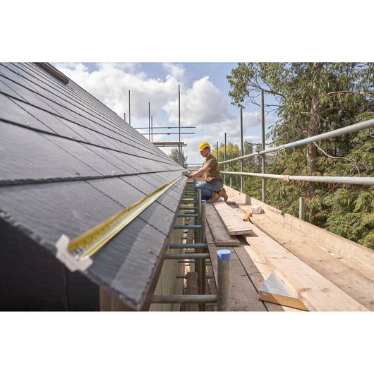 STANLEY FMHT38233-5 tape measure extended along roof edge on scaffolding, worker in hard hat measuring.