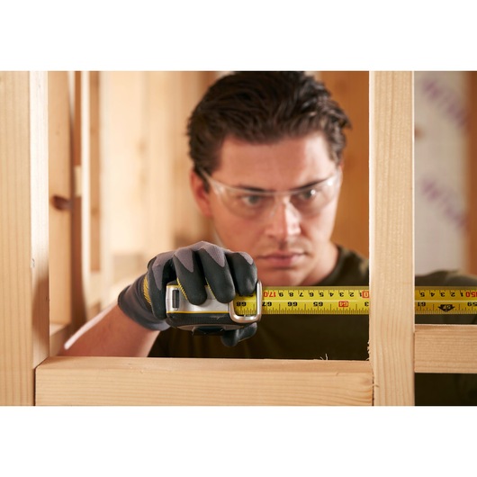 STANLEY FMHT38233-5 tape measure held by gloved hand measuring wooden frame, close-up action photo.