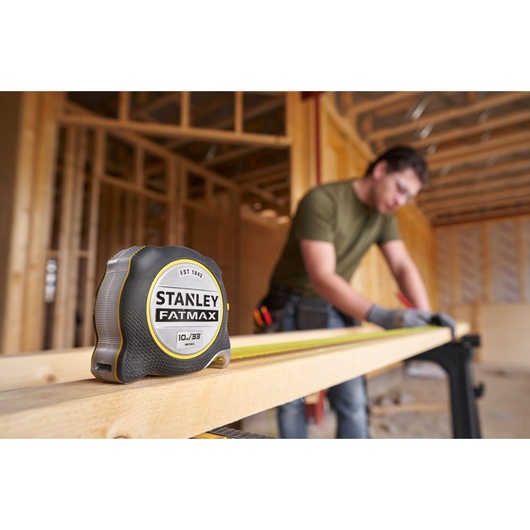 STANLEY FMHT38233-5 FATMAX tape measure on wooden beam with person working in background, construction site setting.