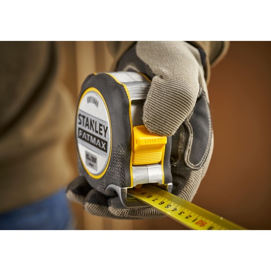 STANLEY FMHT38233-5 tape measure held by gloved hand, yellow and black with clear measurement markings.