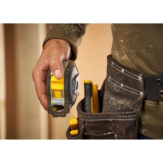 Photo of STANLEY FMHT38233-5 tape measure held in hand next to tool belt with yellow tools.