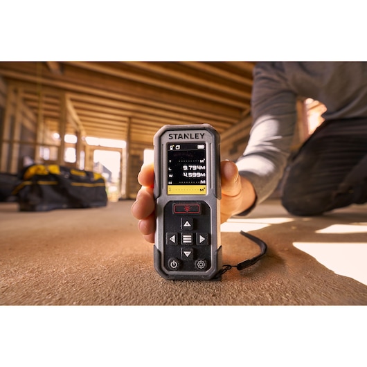 Photo of STANLEY FMHT77050-XJ laser distance measurer being held in a construction setting, display screen visible.