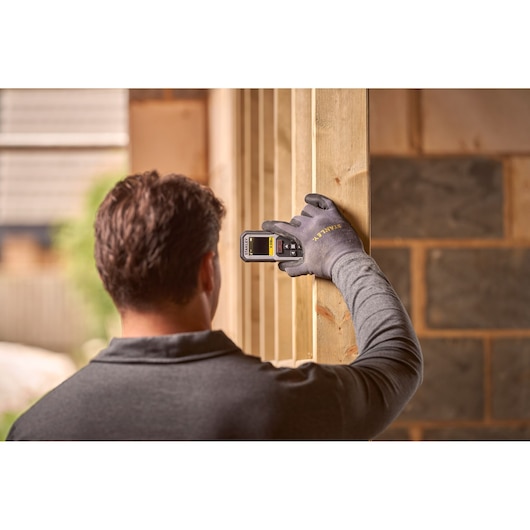 STANLEY FMHT77050-XJ laser distance measurer held by gloved hand against wooden beam, construction site background.