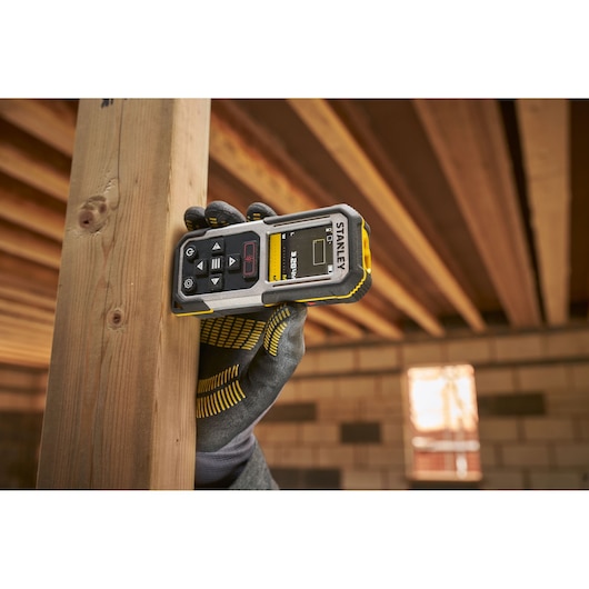 Photo of STANLEY FMHT77050-XJ laser distance measurer in use, showing display and buttons, held in a gloved hand.