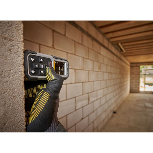 STANLEY FMHT77050-XJ laser distance measurer held by gloved hand against brick wall in construction site.