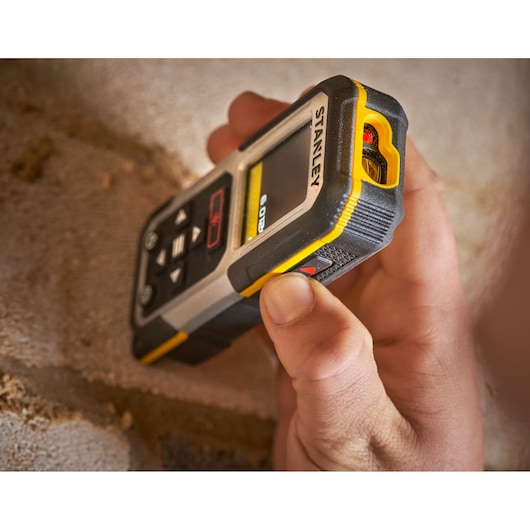 STANLEY FMHT77050-XJ laser distance measurer held against a brick wall, close-up, rugged design.