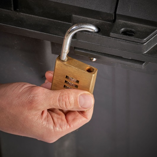 STANLEY FMST17870-1 brass combination padlock held near black storage latch, close-up photo.