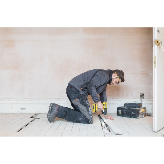 STANLEY SCH20S2K drill in use by worker kneeling, drilling floor planks, toolbox in background, photography.