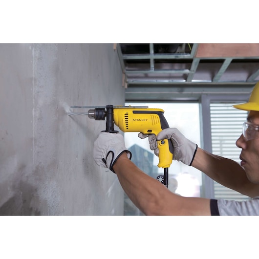 Photo of STANLEY SDH700 yellow corded drill being used on a concrete wall, handled by a gloved worker.