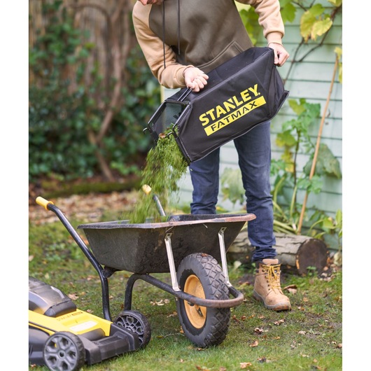 STANLEY SFMCMWS251M lawn mower bag being emptied into wheelbarrow, garden setting, side view.