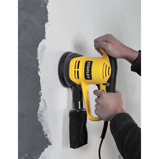 STANLEY SRS480 yellow electric sander in use on a wall, handled by two hands, sanding plaster surface.