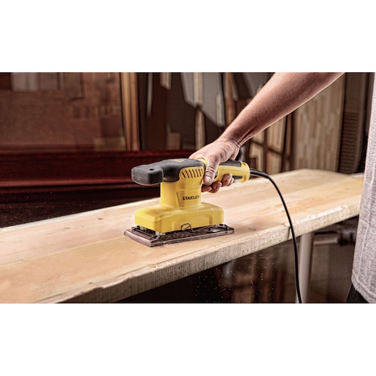 Stanley SS28-AR yellow and black sander in use sanding a wooden plank, close-up photography.