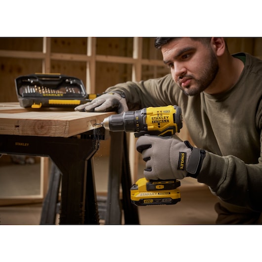 Stanley STA7184 cordless drill in use with drill bit set on wooden workbench, person wearing Stanley gloves.