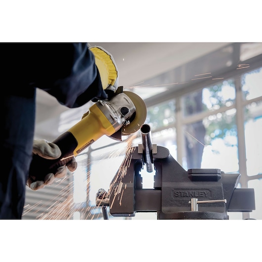 STANLEY STA8061 bench vise holding metal pipe, with sparks flying as worker uses angle grinder.