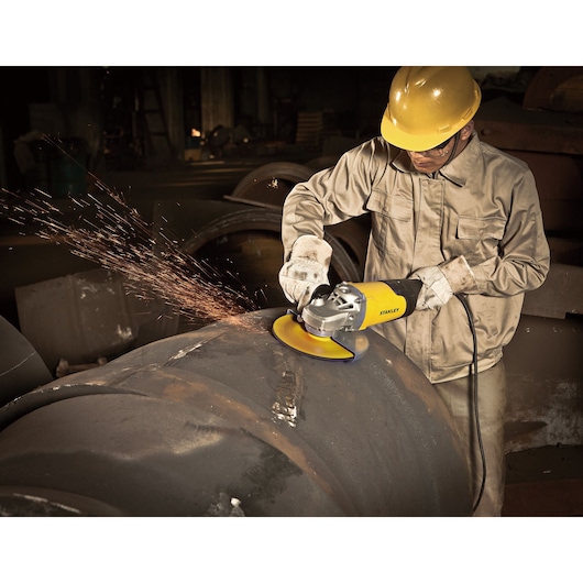 STANLEY STGL2023 angle grinder in use, worker cutting large metal pipe, sparks visible, industrial setting.