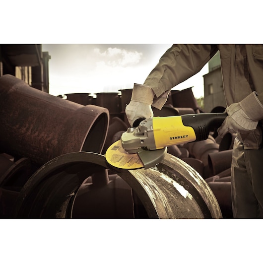 Photo of STANLEY STGL2023 angle grinder used to cut a metal pipe outdoors, showing worker's gloved hands.