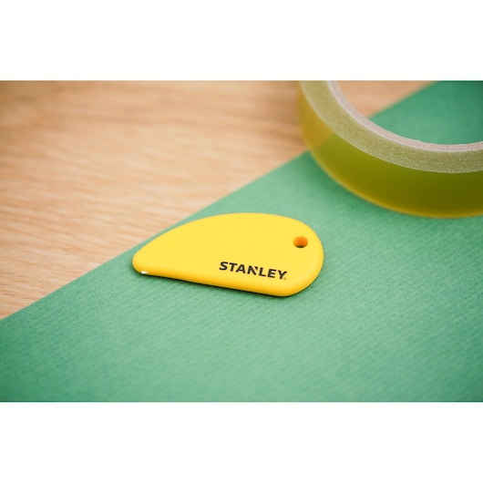STANLEY STHT0-10291 yellow mini safety cutter on green surface, roll of tape in background.