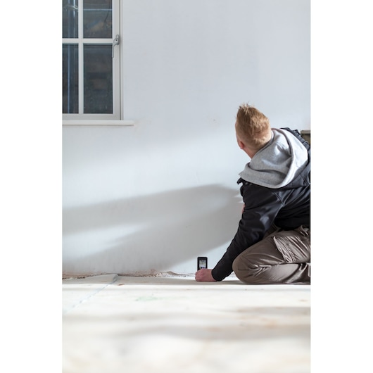 Person using STANLEY STHT1-77139 laser distance measurer on white wall in indoor setting.