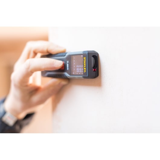 Photo of a hand using STANLEY STHT1-77354 laser distance measurer against a wall, display screen visible.