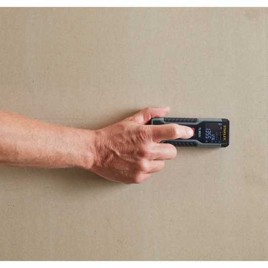 Photo of STANLEY STHT1-77354 laser distance measurer in use on a beige wall, display screen visible.