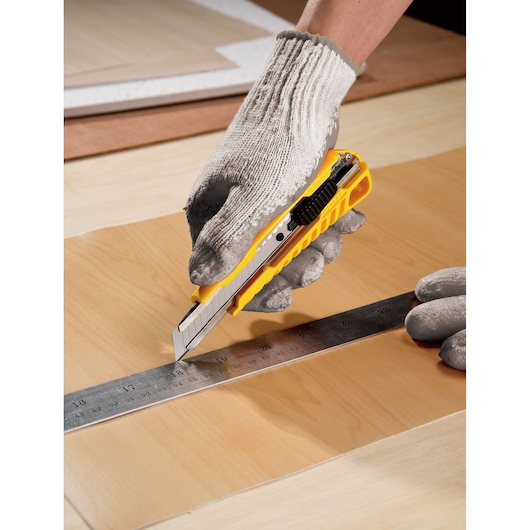 Gloved hand using STANLEY STHT10276-8 utility knife with yellow handle to cut wood along metal ruler.