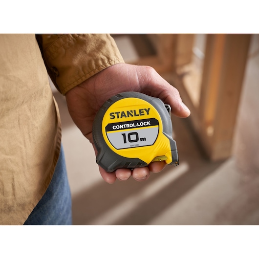 Hand holding a STANLEY Control-Lock 10m tape measure with yellow and gray casing. Indoor construction setting.