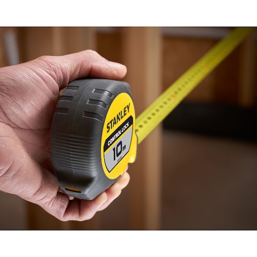 Hand holding a Stanley 10m tape measure with yellow control-lock, tape extended in a construction setting.