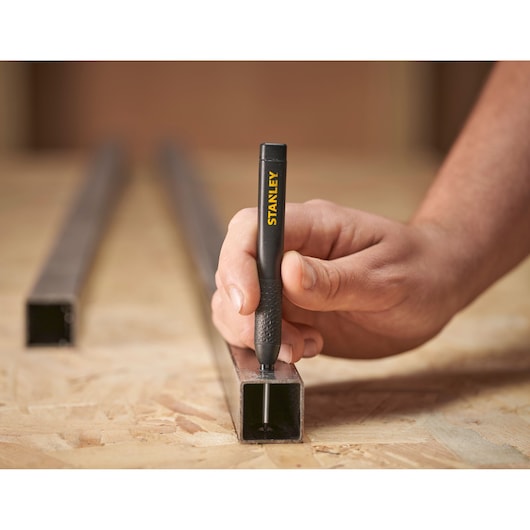 Hand using a Stanley marking pen to write on a metal tube placed on a wooden work surface.