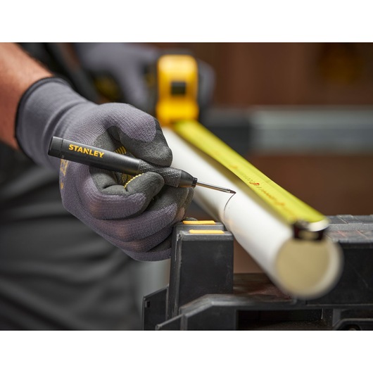 Gloved hand uses a STANLEY marking tool and tape measure to mark a white pipe in a workshop setting.