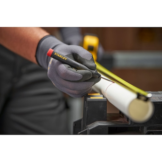 Gloved hand using a STANLEY marker to mark a white pipe with a tape measure on a workbench.