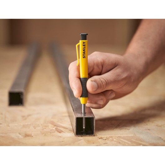 Hand using a STANLEY spring-loaded center punch on a metal square tube placed on a wooden work surface.
