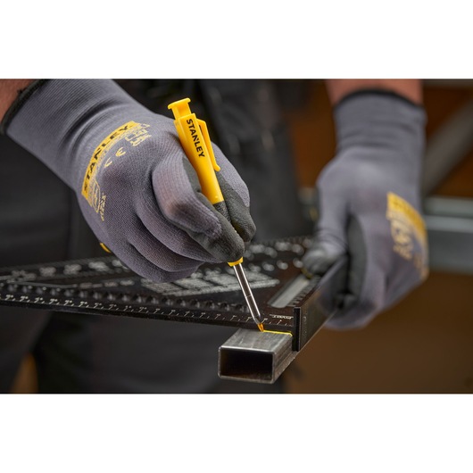 Gloved hands use a yellow STANLEY marking tool with ruler on a metal tube for precise measurement and marking.