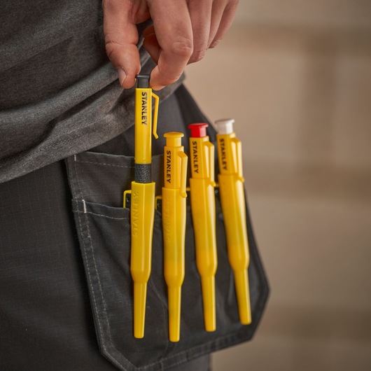 A hand holds a yellow STANLEY tool from a pocket with four yellow STANLEY marking tools clipped to work trousers.
