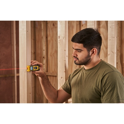 Photo of STANLEY STHT77065-0 laser distance measurer in use, measuring wood studs in a construction setting.