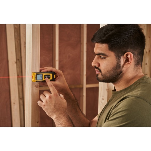 STANLEY STHT77065-0 laser distance measurer in use, showing display and red laser beam in construction setting.